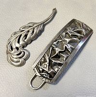 Two antique silver jewelry pieces: an embossed hair barrette clip with crumpled texture and polished frame, and a leaf-shaped reticulated brooch with openwork design.