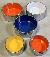 Five stackable chrome ashtrays with colorful enamel interiors (white, orange, blue, yellow) arranged on a flat surface.
