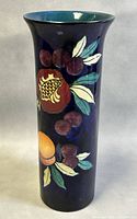 Front and angled view of the 8.5 inch tall dark blue vase decorated with hand-painted pomegranate, berries, and other fruit with green leaves on front, flared rim showing turquoise interior glaze.