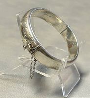 Photo of silver etched floral bangle bracelet with safety chain on stand, showing side profile