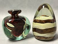 Front view of two Mdina blown glass paperweights: an egg-shaped striped piece and an abstract vase-shaped piece, both signed and with swirled internal colors