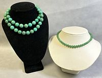 Two necklaces displayed on black and white busts. The shorter light green glass bead choker is on the white bust and the longer turquoise bead necklace is on the black bust.