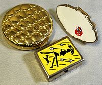 Three vintage items displayed on cloth: a gold basket weave pill box, a white Ladybug Estee Lauder compact, and a Stratton lipstick holder with yellow and black silhouette design and rhinestones.