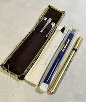 Four vintage writing instruments including a mechanical pencil, two pens, and a ball pen refill laid out on a light surface, two pencils in fabric sleeves.