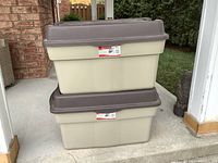 Two stacked Rubbermaid Roughneck storage boxes with beige bases and brown lids on a concrete surface near a brick wall.