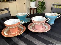 Four vintage Aynsley corset teacups in burnt orange, pink, turquoise, and robin egg blue with three matching saucers on a black tablecloth.
