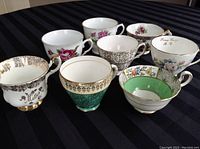 Eight vintage fine china teacups arranged on a black striped fabric background, showing various floral and ornamental designs in different colors including green, pink, gold, and white.