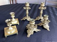 Photo showing the complete set of six brass candlesticks on a dark cloth background, four ornate with detailed bases and two simpler with square bases.