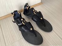 Pair of black leather sandals with gold accent hardware displayed on wood floor.