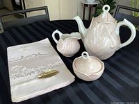Photo of tea set on a black table including teapot with lid, milk jug, sugar bowl with lid, and a folded tablecloth with embroidery