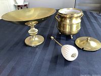 Wide shot showing the footed brass bowl, brass pot with lid and handle, and the large stone stirrer on a dark cloth surface.