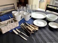 Top view of table showing white Corelle plates, bowls, white mugs, clear drinking and wine glasses, blue placemats, set of steak knives with wood and cream handles, smaller spreader knives, and wicker cutlery basket.