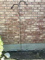 Full view of tall black wrought iron plant stand with two hooks, placed against a brick wall.