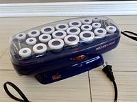 Front view of electric ceramic hair rollers in blue base with closed clear lid, showing white rollers inside and power cord.