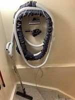 Kenmore central vacuum hose with grey protective sleeve and white power cord hanging on wall along with vacuum handle and wand attached to floor brush.