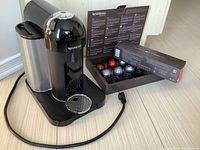 Front angle of black Nespresso Vertuo coffee machine with removable water tank and open box of 18 coffee capsules of various colors behind it on floor.