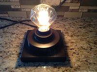 Lamp lit on kitchen counter showing diamond shape clear light bulb and chocolate brown square metal base.