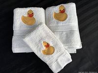 Three white towels displayed, two folded body towels and one hand towel, each with yellow rubber duck embroidery.