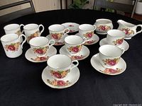 Seven Sadler teacups with eight matching saucers and three coffee mugs arranged on a black cloth, showing floral rose patterns on white ceramic.
