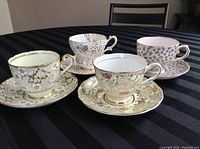 Four vintage teacups with matching saucers displayed on a black striped tablecloth showing their detailed patterns and colors from a front angle.