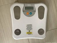 Front view of the white Tanita Ultimate Scale showing the full surface with metallic electrodes and caution sticker.