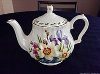 Side view of the white fine china teapot decorated with colorful floral patterns including daffodils, irises, and other flowers, with gold accents on the lid and spout tip.