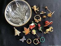 Top-down view of all twelve pins and brooches arranged with the round frosted glass jewelry tray on a black background.