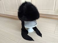 Front angle view of the black muskrat fur hat showing the plush fur and long black fabric ties hanging down.