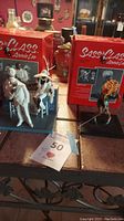 Three figurines displayed with original red Sass 'N Class Annie Lee boxes behind them on a reflective surface: two seated figures and one standing singer.