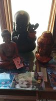 Three Buddha statues placed on glass table with various papers underneath. Dark ceramic bust planter in back holding small cactus, red sitting Buddha in front left, reddish bust with gold swirls front right.