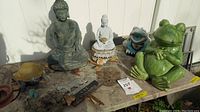 Photo showing two Buddha statues, large green frog planter, smaller blue-green frog figure, turtle figure, and other garden items placed on a weathered outdoor surface.