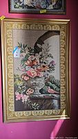 Full view of the framed floral needlepoint with detailed flowers and birds, enclosed in gold gilt wood frame against pink wall backdrop