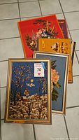 Group photo showing five needlepoint pieces stacked on a tile floor, some framed with silver-colored frames, colors including blue, orange, tan, purple, and red backgrounds