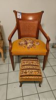 Wood framed needlepoint armchair with matching upholstered foot stool with floral tapestry overcaster wheels, photographed together on tiled floor.