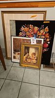 Three framed prints and one unframed needlepoint artwork. Prints include a girl with dog and doll, a floral needlepoint, and other prints in the background.