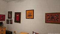 Wide angle photo showing six framed art pieces hanging on a wall including needlepoint and prints.