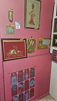View of the corner wall showing various framed textile and brass art pieces including embroidered Asian figure in red dress, winter cabin needlepoint, brass birds, and tiger landscape.