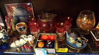 Overview of the assortment of cranberry metal lidded jar, skull cup, glasses, plates, wooden shoes, glass tile, vase, mini tea set, spoons and jars arranged on a shelf.