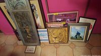 Eleven framed pieces of wall art arranged leaning against a wall, showing a variety of sizes and subjects including a lion, sea captain, and landscape.