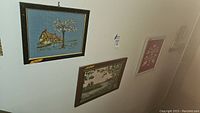 Framed needlepoint art showing a house and tree with a blue sky, hung on wall.