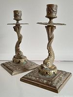 Front angle view of both bronze/brass dolphin candle sticks showing base and candlestick detail with patina.