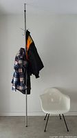 Mid-century modern tension pole coat rack holding several coats, next to a white modern chair for scale.