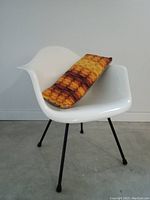 Yellow and brown Mid-Century Modern Op Art geometric patterned rectangular chair cushion placed on a modern white molded plastic chair with metal legs.
