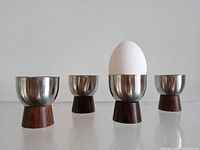 Four vintage Danish egg cups displayed in a line with one holding a white egg, showing reflective stainless steel cups and dark rosewood bases.