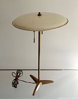 Full view of lamp showing white saucer shade, slender stem, tripod base and attached cord