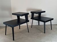 Pair of vintage two-tier side tables with black vinyl tops and angled tapered legs, viewed front angle.