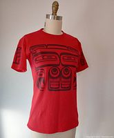 Front and left side view of the red shirt showcasing large black Haida art motif across the chest and on sleeves.