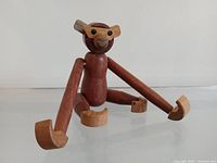 Front view of the wooden monkey toy showing red teak body, lighter wood ears, hands, and feet, with black eyes and rounded snout.
