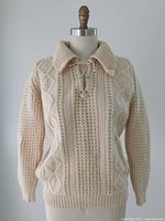Front view of cream-colored Aran wool sweater on mannequin showing cable knit and lace-up collar detail.