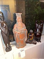 Photo showing four figurines including two tall African figures, an Asian robed figure, and a small seated Buddha figure with a yellow beaded necklace, displayed on a windowsill.